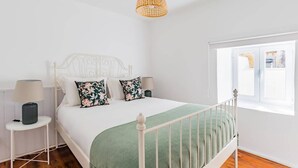 Una cama de metal blanco con colcha verde, dos mesitas de noche con lámparas y una lámpara colgante de mimbre.