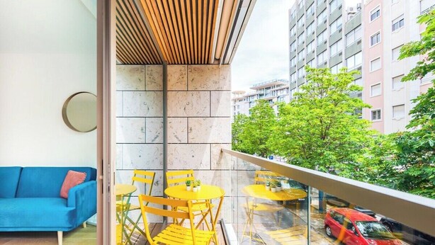 Un balcon avec des chaises jaunes et un canapé bleu, un mur carrelé et une vue sur une voiture rouge et des immeubles.