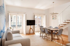 A living room with a sofa, a TV on a tripod, a dining table with chairs, and a staircase. - Baixa Rooftop Duplex | Lisbon (Lisbon)