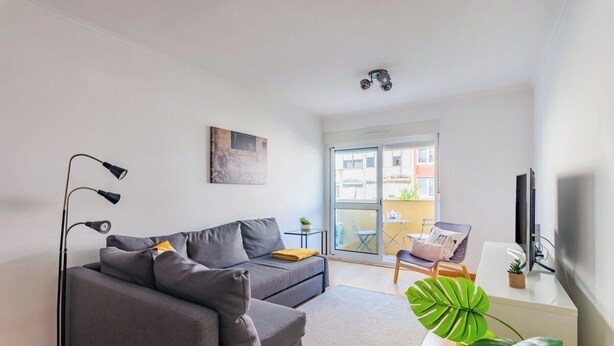 A modern living room with a sofa, a floor lamp, a TV, and a large window. - Olaias Park Flat (Lisbon)