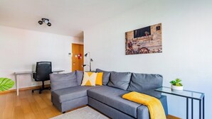 A modern living room with a grey sectional sofa, a glass coffee table, a small desk with a chair, and a wall-mounted lamp. - Olaias Park Flat (Lisbon)