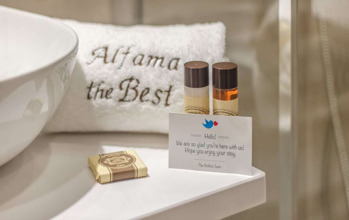 Ein Badezimmer mit einem weißen Waschbecken, einem Handtuch mit dem Aufdruck 'Alfama the Best', zwei Flaschen auf einem Regal und einer Karte mit einer Nachricht.