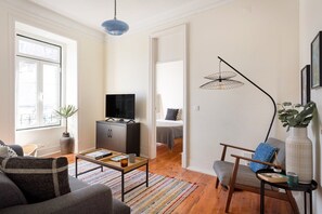Ein Wohnzimmer mit einer Couch, einem Couchtisch, einem Fernseher auf einem Schrank, einer Stehlampe und Blick durch eine offene Tür.