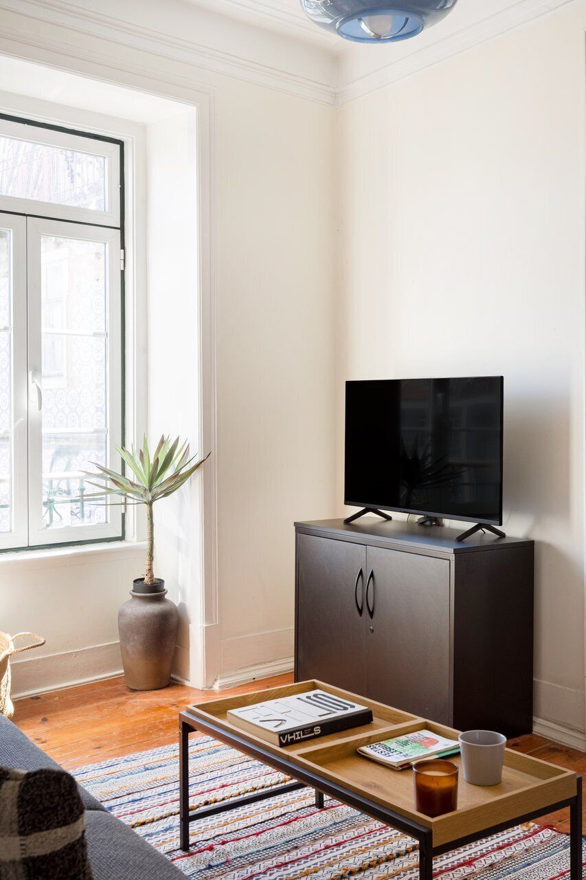 En moderne stue med et fladskærms-tv på et skab, et sofabord med en bog og en kop, en potteplante og et stort vindue.