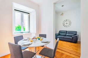 Ein Essbereich mit einem runden Tisch, grauen Stühlen und Frühstücksangeboten. Ein schwarzes Sofa steht an der Wand.