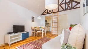 A living room with a TV, white sofa, wooden floor, and a dining area with chairs and a table. - Bright Alfama Loft with Mezzanine | Guest Inn (Lisbon)