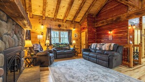 A log cabin interior with a fireplace, two leather sofas, a patterned rug, and a stone wall.