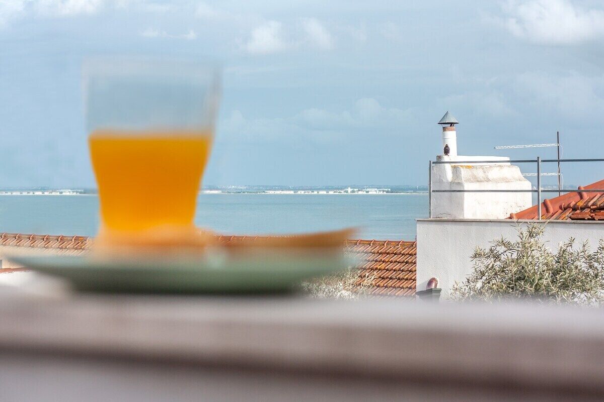 A glass of orange juice on a plate, a white building with a chimney, and a view of the sea.