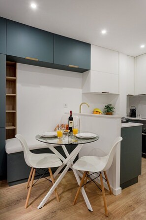 A modern kitchen with white cabinets, a dining area with two white chairs, a glass table, and a small island with a sink and fruit. - Petite Charm in Old Lisbon (Lisbon)