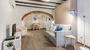 Ein modernes Wohnzimmer mit einer Couch, einem kleinen Tisch mit Lampe, einem Fernseher und einem Schreibtisch mit Computer.