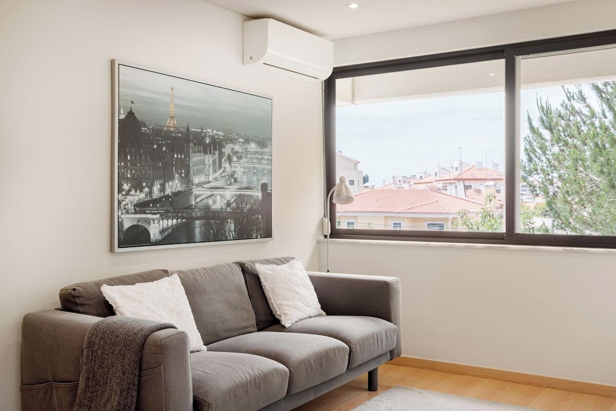 A modern living room with a grey sofa, a large window with a view of buildings, and a framed cityscape picture on the wall.