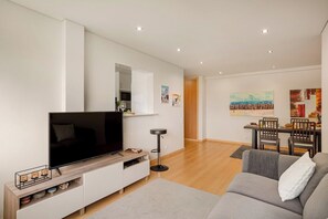 Ein modernes Wohnzimmer mit Flachbildfernseher, einem grauen Sofa, einem Essbereich mit Holztisch und Stühlen sowie einem Flur, der zu einem weiteren Raum führt.
