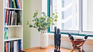 Una habitación con un escritorio de madera, una estantería blanca llena de libros, una planta en maceta y una ventana con vistas a edificios.