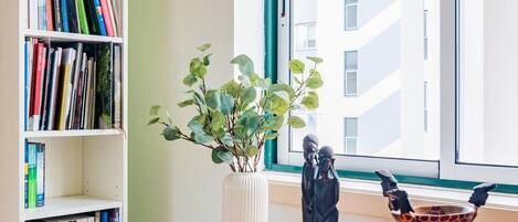 A room with a wooden desk, a white bookshelf filled with books, a potted plant, and a window with a view of buildings.