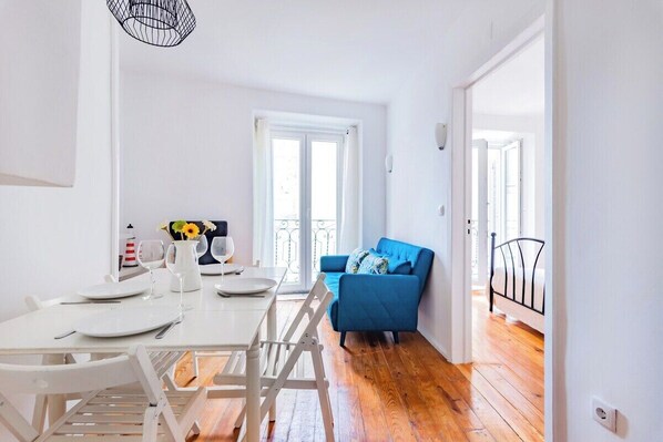 Ein helles Zimmer mit einem Esstisch für zwei Personen, einem blauen Sofa und einem Bett mit Metallgestell.