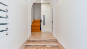 A hallway with a wooden staircase leading to a door, white walls, and a metal rack on the left. - Conde 3BDR Luxury with Balcony (Lisbon)