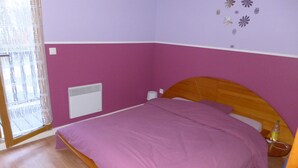 A bedroom with a bed, a window with curtains, a wall-mounted heater, and a bottle on the nightstand.