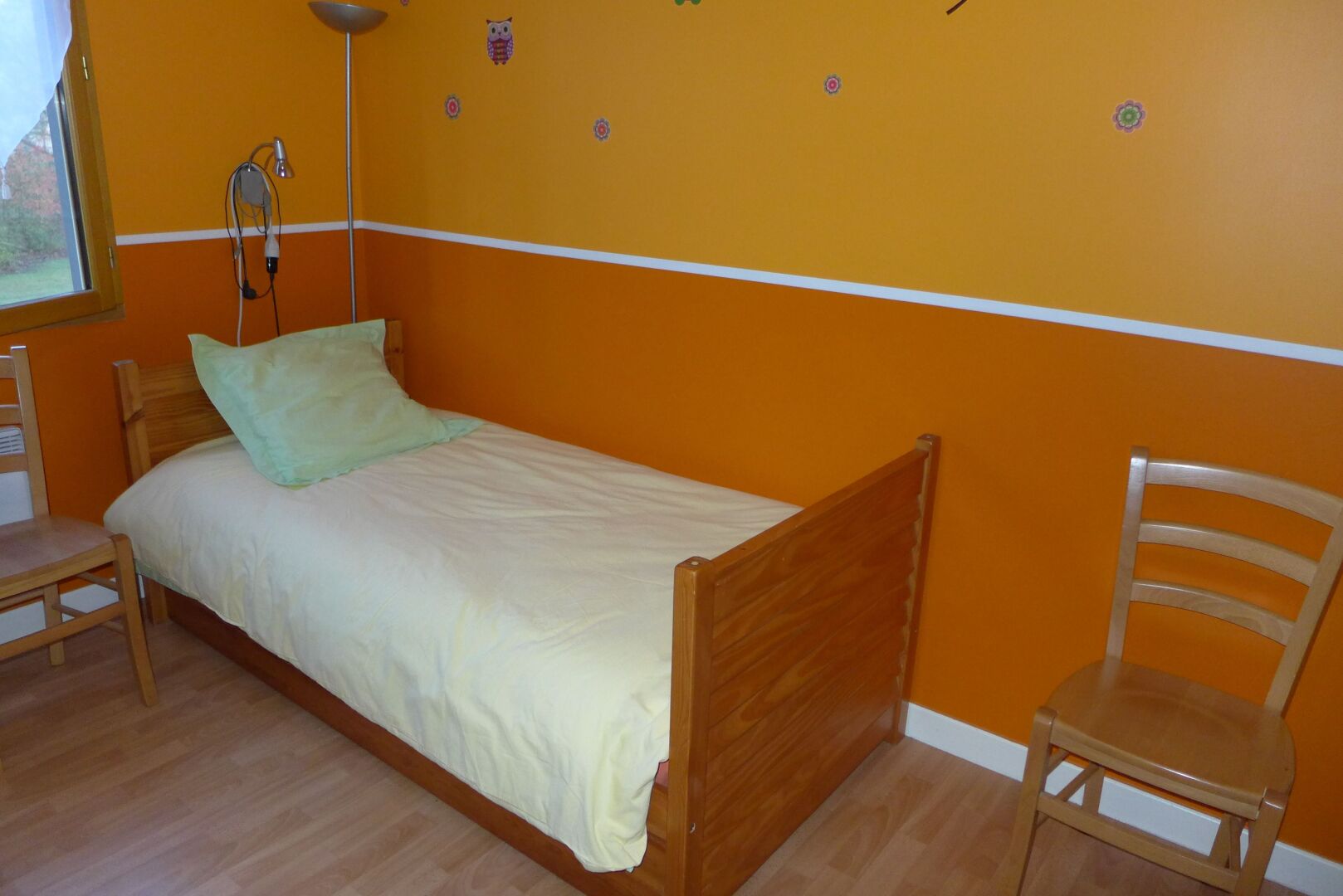 Una cama individual con estructura de madera y un almohadón verde, una silla de madera y una lámpara sobre un suelo de madera en una habitación con paredes anaranjadas.