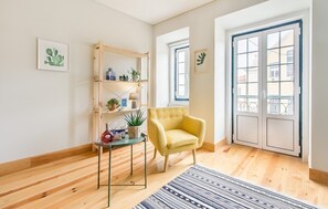 Ein Zimmer mit Holzboden, einem gelben Sessel, einem Holzregal mit Pflanzen, einer gerahmten Kakteenkunst und einem großen Fenster mit Ausblick.