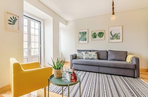 Ein Wohnzimmer mit einer Couch, einem gelben Sessel, einem kleinen Tisch mit Pflanze und Flasche sowie gerahmten Bildern an der Wand.