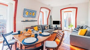 Ein Essbereich mit Holztisch, Stühlen und einer Frühstücksecke. Im Hintergrund befindet sich eine Couch und ein Fernseher.