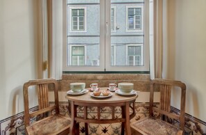 Ein Eckzimmer mit Fensterblick auf ein Gebäude, ein Holztisch mit Tellern und Tassen sowie zwei Holzstühle.