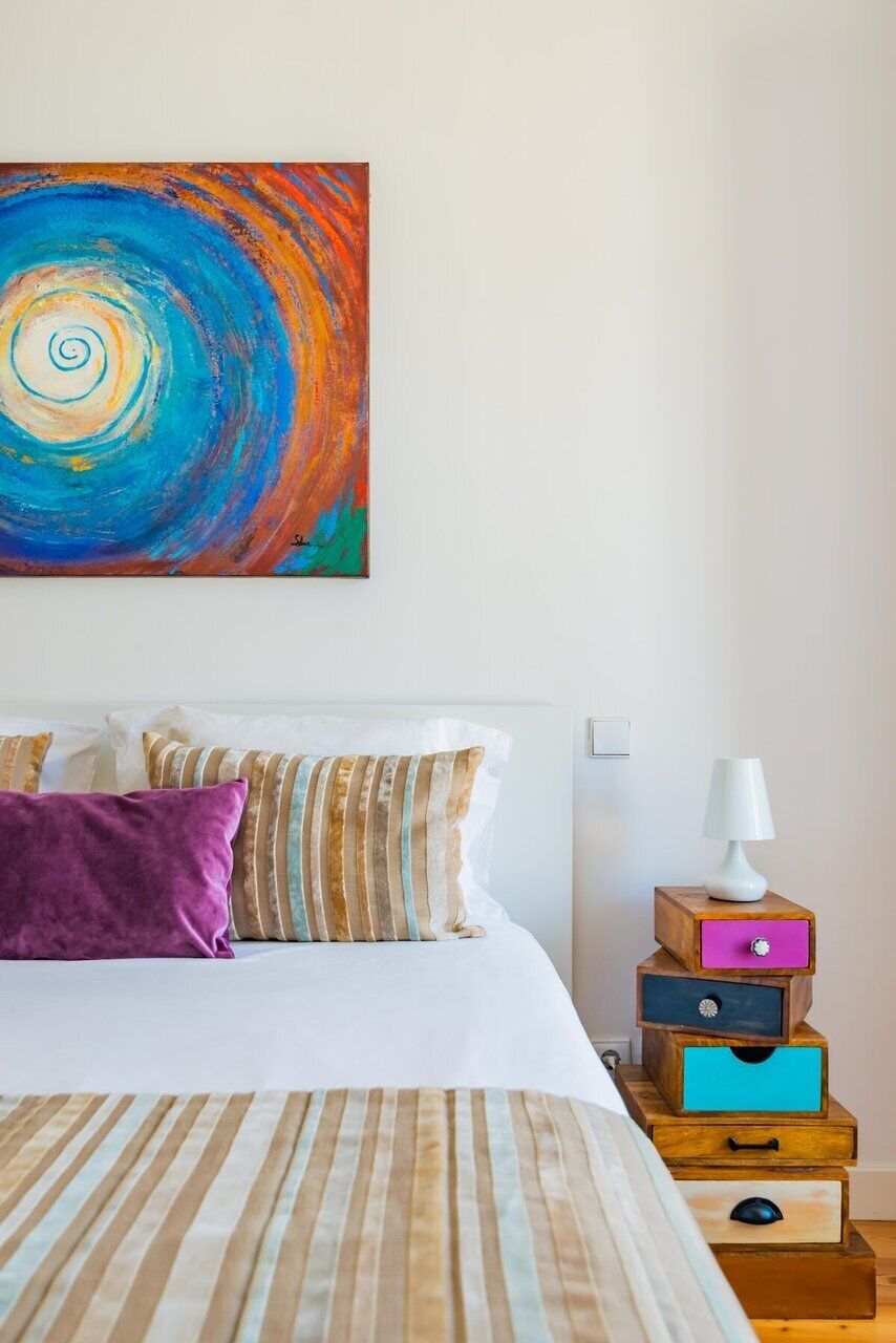 A neatly arranged hotel room with a bed, pillows, a bedside table with a lamp, and a colorful abstract painting on the wall.