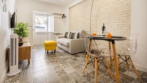 Un espacio habitable pequeño con sofá, una mesa de comedor con sillas, un taburete, una planta en maceta y un televisor montado en la pared.