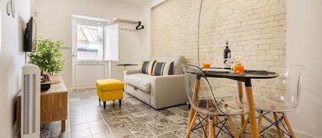 A compact living space with a sofa, a dining table with chairs, a small stool, a potted plant, and a wall-mounted TV.