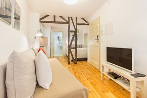 Ein Zimmer mit Holzboden, einem weißen Sofa mit Kissen, einem Fernseher auf einem Schrank und einer Treppe, die in ein weiteres Zimmer führt.
