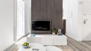 Ett modernt vardagsrum med en platt-TV som är monterad på en mörk träpanelvägg, ett vitt konsolbord med dekorativa föremål och ett runt soffbord med en skål med frukt och en bok.