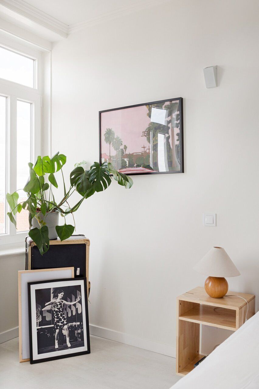 Ein Zimmer mit Fenster, einer Topfpflanze, einem gerahmten Bild, einer Lampe und einem kleinen Holztisch.