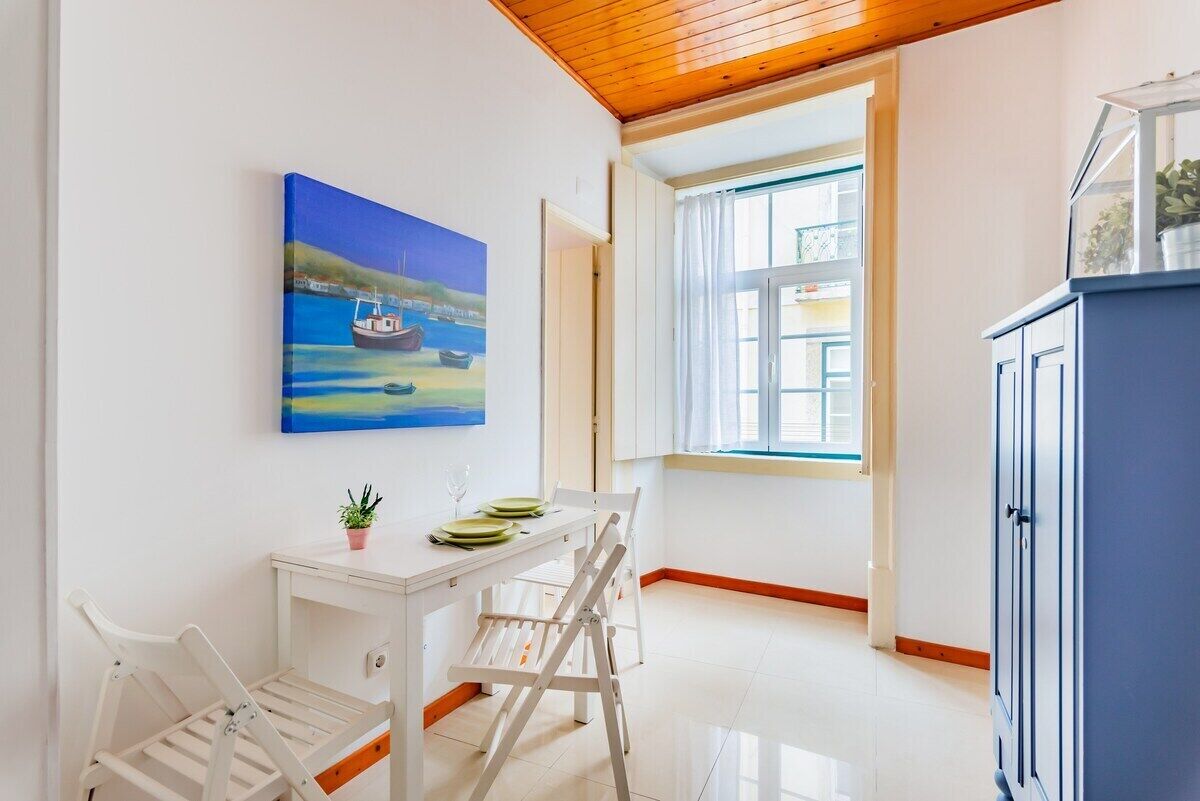Een kamer met een witte tafel, stoelen, een schilderij van boten, een kast en een raam met gordijnen.