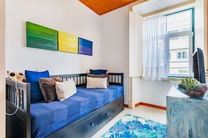 Um quarto com uma cama, uma janela com cortinas, uma escrivaninha com uma planta e uma parede com três quadros.
