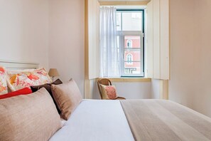 Ein Hotelzimmer mit einem Bett, Kissen, einem Sessel, einem Fenster mit Vorhängen und Blick auf ein Gebäude.