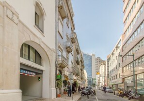 Eine schmale Stadtstraße mit mehrstöckigen Gebäuden, einem sichtbaren Hauseingang mit einem grünen Schild und abgestellten Rollern.