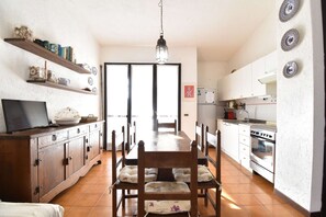 A dining area with a wooden table and chairs, a television on a cabinet, and a kitchen with white cabinets and a stove. - Cottage with fenced garden and covered veranda (Aglientu)