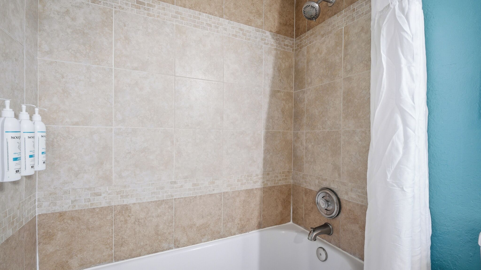 Un baño con baldosas color beige, una bañera blanca, un cabezal de ducha y tres botellas de jabón en la pared.