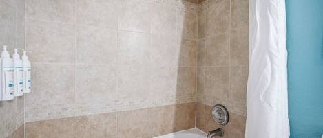 Un baño con baldosas color beige, una bañera blanca, un cabezal de ducha y tres botellas de jabón en la pared.