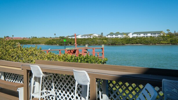 A wooden deck with white chairs and a red railing overlooking a calm body of water, with buildings in the background. - Intracoastal Townhome with Private Entrance, Beach Equipment, and Bikes! (Indian Shores)