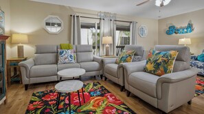 Ein Wohnzimmer mit zwei grauen Sofas, einem kleinen runden Tisch, einem farbenfrohen Teppich mit Blumenmuster und einem Schild mit der Aufschrift 'Beach House'.