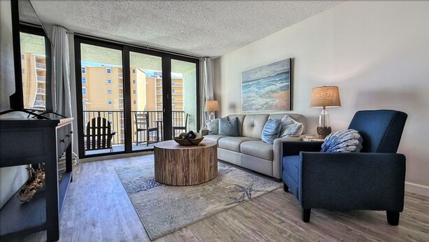 A modern living room with a sofa, armchairs, a coffee table, and a view of a building through large windows. - Quintessential Beachfront Condo escape right on the Sand with private balcony (New Smyrna Beach)