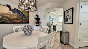 A dining area with a white table and chairs, a decorative vase, a chandelier, and a painting of a bird. - Quintessential Beachfront Condo escape right on the Sand with private balcony (New Smyrna Beach)