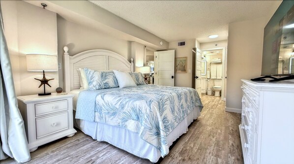 A bedroom with a bed, nightstands, a dresser, and a bathroom visible through an open door. - Quintessential Beachfront Condo escape right on the Sand with private balcony (New Smyrna Beach)