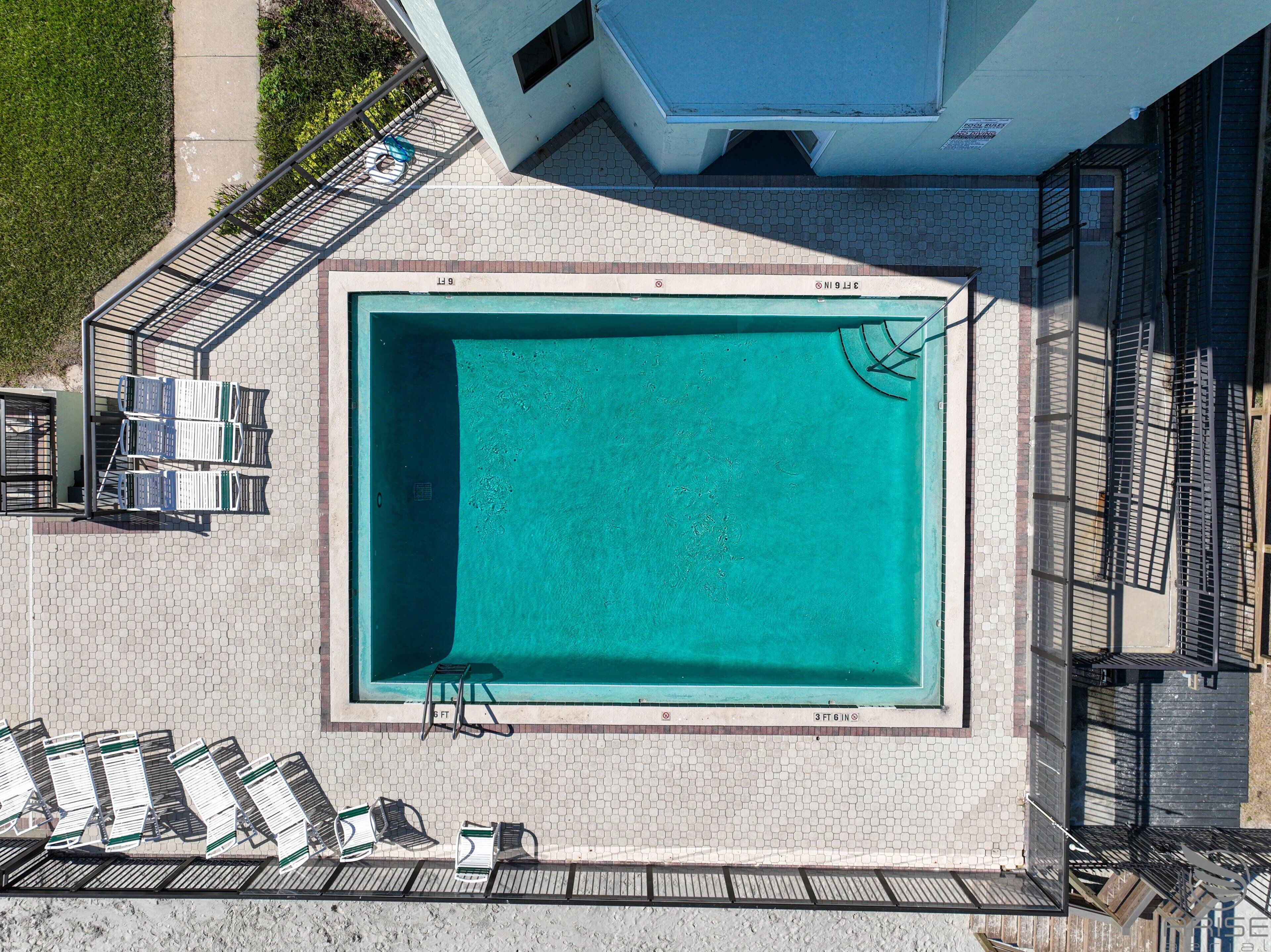 An aerial view of a rectangular swimming pool with a tiled deck surrounding it, equipped with lounge chairs and a sunbed.