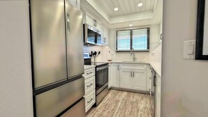 A compact kitchen with white cabinets, a stainless steel refrigerator, and a window with blinds. - Quintessential Beachfront Condo escape right on the Sand with private balcony (New Smyrna Beach)