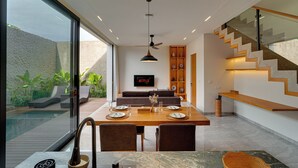 A modern dining area with a wooden table set for four, a sofa in the background, and a staircase with glass railings. - Villa Vibhava - Umalas (Seminyak)