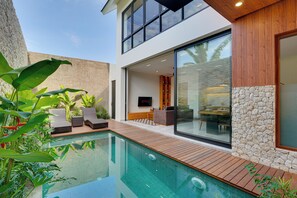 A modern house with a swimming pool, wooden deck, and a large glass door leading to a living area with a TV. - Villa Vibhava - Umalas (Seminyak)