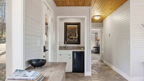 A narrow hallway with white walls and a wooden ceiling, leading to a kitchen area with white cabinets, a black dishwasher, and a stone floor. - Little House on Hickory (Lowell)
