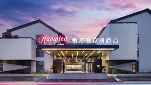 Un hotel moderno con un llamativo letrero de 'Hampton by Hilton', que cuenta con una entrada de cristal y un área exterior cubierta.